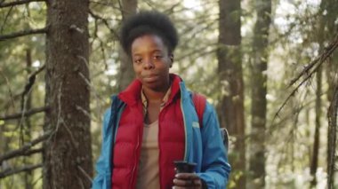 Genç siyahi kadın turistin belden aşağısı, yürüyüş direkleri ve sırt çantasıyla, yaz günü ormanda kameraya poz verirken çekilmiş.