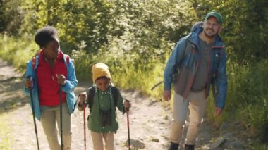 Mutlu çoklu etnik ebeveynler ve küçük melez oğul yaz günü yürüyüş direkleri ve sırt çantalarıyla ormanda yürürken gülümsüyor ve konuşuyorlar.