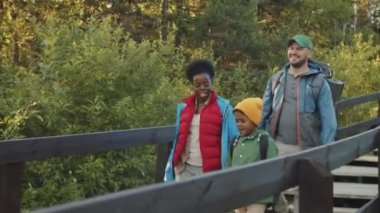 Pozitif Afro-Amerikalı anne, beyaz baba ve heyecanlı karışık ırk oğlu ulusal parktaki tahta merdivenlerde sırt çantalarıyla yürüyor, yaz günü yürüyüş yaparken gülümsüyor ve sohbet ediyorlar.