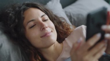 Genç ve güzel bir kadın, sabah uyandıktan sonra evde yatıp gülümsüyor ve akıllı telefondan mesaj yazıyor.