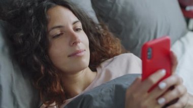 Kafkas 'lı genç bir kadının yatağında yatarken ve akıllı telefonuyla cep telefonuna bakarken yüksek açılı çekimini kapat.