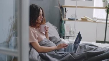 Genç bir kadın sabah uyandıktan sonra evinde oturuyor, fincandan kahve içiyor ve laptopunu kullanıyor.