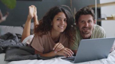 Genç ve neşeli çiftin portresi dizüstü bilgisayarla yatakta uzanıyor, kameraya bakıyor ve gülümsüyor.