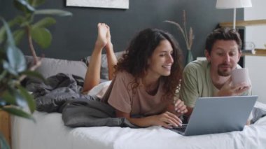 Genç ve güzel bir çift yatakta birlikte uzanıyor, kahve içiyor, internette dizüstü bilgisayarda geziniyor, sohbet ediyor ve gülümsüyor.