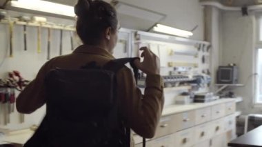 Genç beyaz erkek marangozun atölyesine girerken belden yukarısından çekilen fotoğrafı. Işığı açıyor, sırt çantasını ve ceketini çıkarıyor, masaya bırakıyor ve duvar rafından bazı aletleri alıyor.