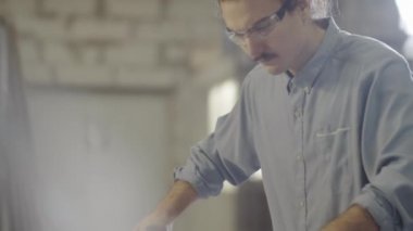 Güvenlik gözlükleri takmış genç beyaz marangozun ahşap testeresinde tahtayı itme çubuklarıyla hareket ettirirken dikkatli bir şekilde odun keserken çekilen görüntüsü.