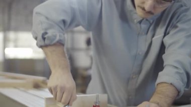 Güvenlik gözlüklü genç beyaz marangozun yakın plan çekimini indiriyorum. Tahtayı kaydırma masasına yerleştiriyorum. Kesmek için vidaları vidalamış kıskaçlarla kesiyorum.