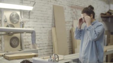 Beyaz tenli genç marangozun atölyede durup güvenlik gözlüğü taktığı, büyük dolap testeresini açıp açtığı ve tahta parçasını kaydırma masasına kelepçelemeye başladığı kavis çekimi.