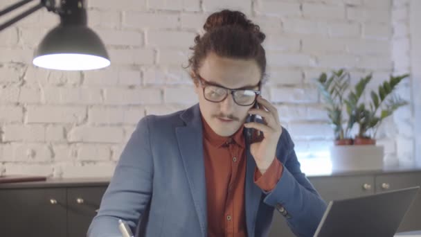 戴眼镜 留着胡子和男人发髻的白人男性创业者坐在阁楼办公室的办公桌前 在手机上进行商务对话 用笔写下信息的勾心斗角 — 图库视频影像