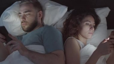 Genç beyaz erkek ve kadının gece yatakta birlikte yatarken, ellerinde akıllı telefonlar, sosyal ağlardaki haber kaynaklarını incelerken ve incelerken göğüsten çekilen görüntüleri.
