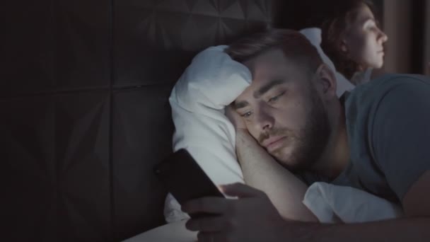 照片中年轻的白人男子深夜躺在床上 带着迷人的神气拿着智能手机 翻阅着新闻资料 妻子则忙着拿着身边的手机 — 图库视频影像