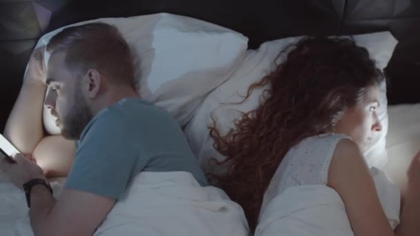 白种人年轻的丈夫和妻子晚上躺在床上 背对背地躺在床上 全神贯注地使用他们的智能手机 这让人神魂颠倒 — 图库视频影像