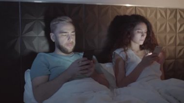 Akşamları pijamayla yatakta uzanmış, yatak başlığına yaslanmış, tamamen akıllı telefonlara dalmış, dikkati dağılmış beyaz erkek ve kadının belden yukarı pozu.