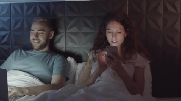 ノートパソコンやスマートフォンを使い笑顔で何かを話しながらおしゃべりをしている若い白人男性と女性がベッドの上に寝そべっている — ストック動画