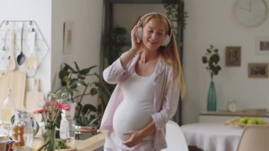 Kablosuz kulaklıklarla mutlu hamile kadın mutfaktaki müzik eşliğinde gülüyor ve dans ediyor.