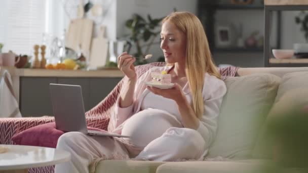陽気妊娠中の女性座っていますソファに自宅でラップトップで彼女の膝の上に デザートを食べます オンライン映画や笑顔を見ます — ストック動画