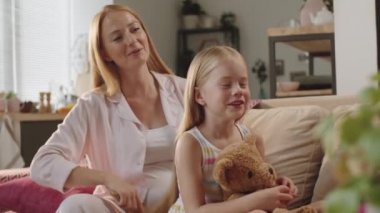 Kanepede oyuncak ayıyla oturan, uzun sarı saçlarını tararken hamile annesiyle gülümseyen ve konuşan tatlı küçük bir kız.