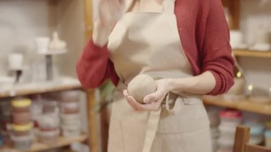 Atölyede duran, elinde kilden bir top tutan, elleri sıvazlayan ve el yordamıyla döven, süveter ve önlükle tanınmayan kadın çömlekçinin ellerinin yakın plan çekimi.