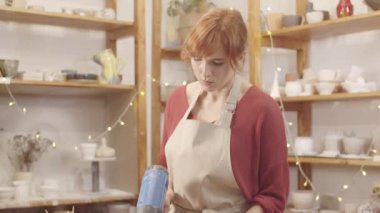 Önlüklü genç beyaz kadın çömlekçinin, atölyede masasında fön makinesini tutarken, çanak çömlek üzerinde ıslak toprak kabını döndürürken ve yüzeyi kururken orta ölçekli bir çekim.