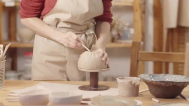 Orta boy el ve orta boy çanak çömlek sanatçısı atölyede ayakta duruyor, çanak çömlek üzerinde el yapımı toprak çanağı kasesi dönüyor ve alttaki kilden tel uçlu aleti kesiyor.