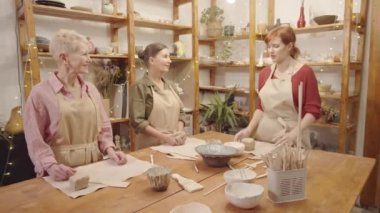Tam kavisli genç beyaz kadın zanaat öğretmeni atölyede iki son sınıf bayan için ustalık dersi veriyor seramik kaplar ve mallar gösteriyor ve çömlek yapımının temellerini anlatıyor.