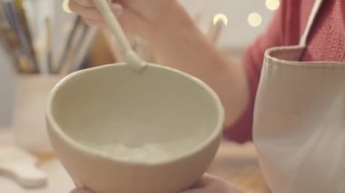 Önlük üzerinde tanınmayan kadın çömlekçinin elleri ile yakın plan çekimde el yapımı toprak kaplarının etrafına sürtünerek kaygan kil yayıyor.