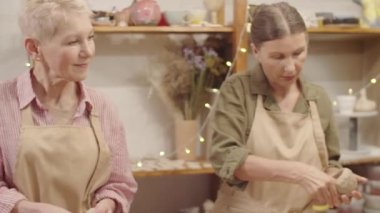 Sanat atölyesinde çömlekçilik dersi sırasında plastik çamurdan kap yapan iki yaşlı beyaz kadın ve doğru tekniği gösteren ve açıklayan genç bayan öğretmen.