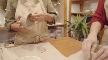 Kimliği belirsiz genç ve iki yaşlı kadının el yapımı kil kaplar yaparak çömlek atölyesindeki plastik kil kaplamalarını yakından çekiyoruz.