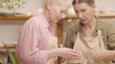 İki yaşlı beyaz kadının seramik atölyesinde kil kaseleri yaparken, bir kadının kenarları yontup tavsiye verirken, arkadaşının da kaburga aletini alıp teknenin dibini düzleştirirken göğsünden çekilen fotoğrafı.