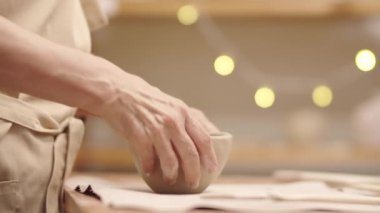 Bej önlüklü, kilden kase yapan, duvarları yontan ve baş parmağıyla dış yüzeyi düzleştiren yaşlı bir bayanın kırış kırış elleriyle yakın çekim.