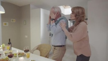 Neşeli son sınıf öğrencisi kadın, kocası elleriyle gözlerini kapatırken kocasıyla masaya doğru yürüyor. Ona hediye paketi veriyor, dilek diliyor ve kadeh tokuşturup kadeh kaldırıyor.
