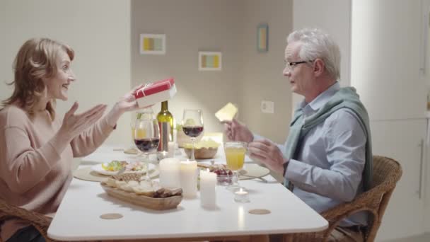 希望を述べ 自宅での休日の夕食時に夫にプレゼントを与えるシニア女性のアークショット 幸せなカップル笑顔とクリンクグラスでワイン — ストック動画