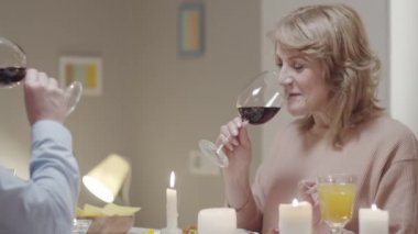 Neşeli bir eş ve koca gülümseyerek ve konuşarak, kadeh tokuşturarak, kırmızı şarap içerek ve evde bayram yemeğinin tadını çıkararak.