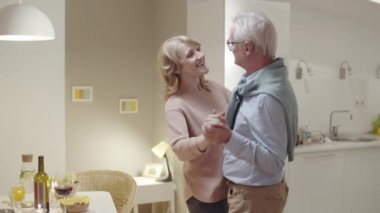 Mutlu yaşlı çift yemek masasının yanındaki salonda dans ederken sohbet ediyor ve romantik anın tadını çıkarıyor.