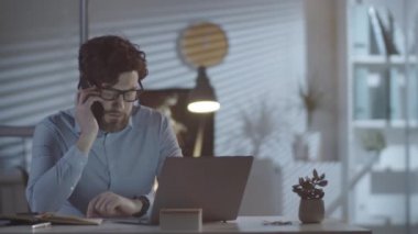 Orta boy sakallı bir adam, karanlık ofisinde masasında oturuyor ve müşterisiyle konuşuyor.