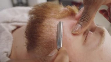 Tanımlanamayan Afro-Amerikan berberin elleri berber dükkanında düz kenarlı jiletle beyaz adamın sakalını kesiyor.