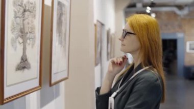 Duvara doğru yürüyen gözlüklü kızıl saçlı kadının yan görüntüsü. Galeride çağdaş sanat sergisini ziyaret ederken resme yakından bakıyor.