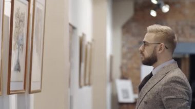 Beyaz tenli gözlüklü ve zarif kıyafetli bir adam sanat galerisinde el ele tutuşmuş, duvardaki sanat eserlerine bakıyor ve bunu anlamaya çalışıyor.