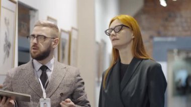 Kadın ziyaretçilere çağdaş sanat koleksiyonunu anlatırken elinde pano tutan ve duvardaki sanat eserlerine işaret eden profesyonel erkek galeri rehberi.