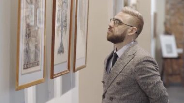 Gözlüklü ve zarif kıyafetli zeki beyaz adam müzede duruyor ve duvardaki sanat eserlerini inceliyor.