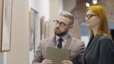 Erkek galeri rehberi panoyu tutuyor, duvardaki resme bakıyor ve modern sanat koleksiyonunu sunarken kızıl saçlı kadınla tartışıyor.