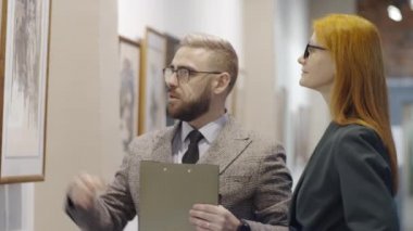 Kızıl saçlı kadına sanat eserlerinden bahseden ve modern sanat koleksiyonunu sunan deneyimli bir erkek galeri rehberi.