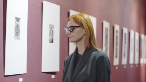 Tänksam Rödhårig Kvinna Glasögon Tittar Konstverk Galleriväggen Och Beundrar Dem — Stockvideo