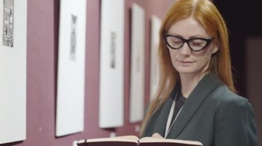 Gözlüklü çekici kızıl saçlı kadın sanat eleştirmeni galeri duvarındaki sanat eserlerine bakıyor ve sergi eleştirisi üzerinde çalışırken not defterine bir şeyler yazıyor.
