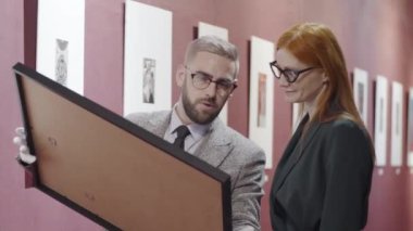 Beyaz pamuk eldivenli erkek sanat uzmanı çerçeveli resim tutuyor ve bunu sergi salonunda birlikte çalışırken bayan galeri müdürüyle tartışıyor.
