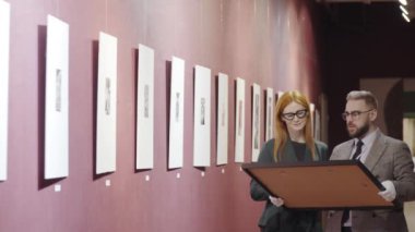 Kadın galeri müdürü duvara işaret ediyor ve beyaz pamuklu eldivenle çerçeveli resmi tutarken erkek meslektaşıyla nereye resim asacağını konuşuyor.