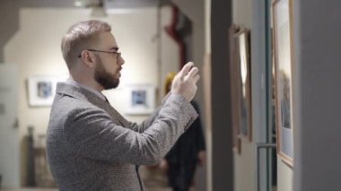 Gözlüklü zarif beyaz adam galeriyi ziyaret ederken duvarda akıllı telefonuyla resim çekiyor.