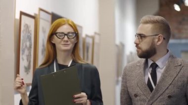 Çekici bayan galeri rehberi elinde bir pano, erkek ziyaretçiyle duvardaki modern çizimleri tartışıyor ve ona çağdaş sanat koleksiyonunu gösteriyor.