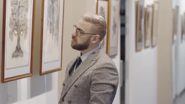 一个戴眼镜 穿着雅致西服的英俊男子站在美术展览馆里 细看墙上的图画 — 图库视频影像