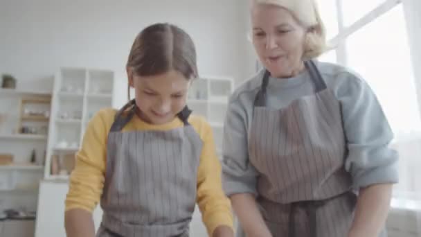 傾きダウンアップショットの陽気な女の子でエプロンロールアウト生地とともに木製ピンでキッチンテーブルの上に準備中クッキーと祖母 — ストック動画